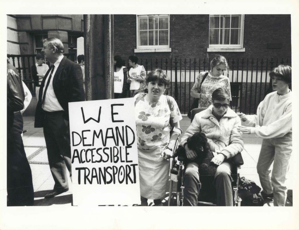 A black-and-white protest photograph with a hand-lettered sign reading We Demand Accessible Transport
